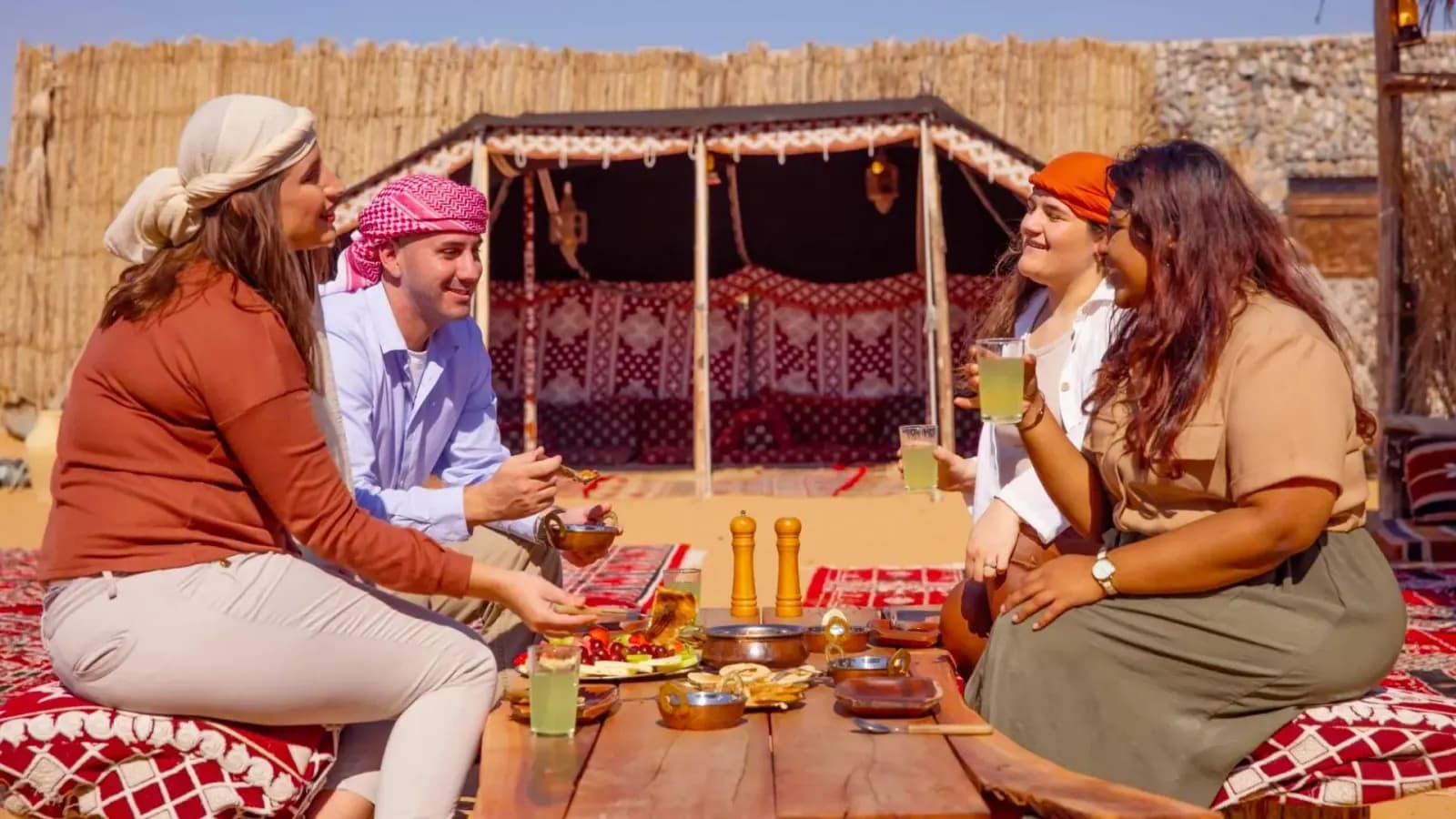 Four travelers sharing drinks and a meal at a desert Bedouin-style camp