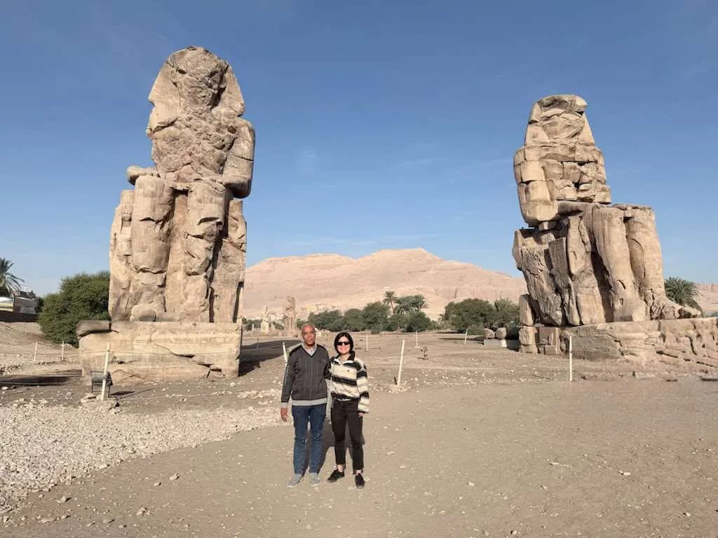 A couple posing between the Colossi of Memnon under a clear blue sky, Luxor